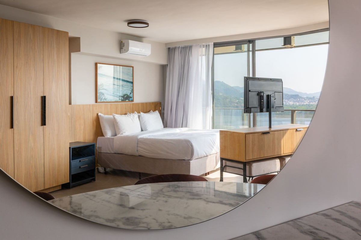A modern hotel room with a bed, a desk, a TV, and a large window offering a view of the city.
