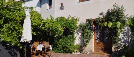 A courtyard with a table and chairs, surrounded by greenery and a building with a tiled roof.
