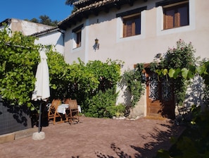 A courtyard with a table and chairs, surrounded by greenery and a building with a tiled roof.