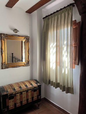 A room with a large ornate mirror, a bench with a patchwork design, and a window with a sheer curtain.