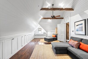 A spacious attic room with a sloped ceiling, wooden beams, a ceiling fan, a gray sofa with orange cushions, a small table, and a framed picture on the wall. - 1868 R. H. Wood House | Historic Rockport Retreat (Rockport)