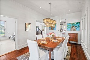 A dining room with a wooden table set for six, white chairs, a chandelier, and a painting of a palm tree. - 1868 R. H. Wood House | Historic Rockport Retreat (Rockport)