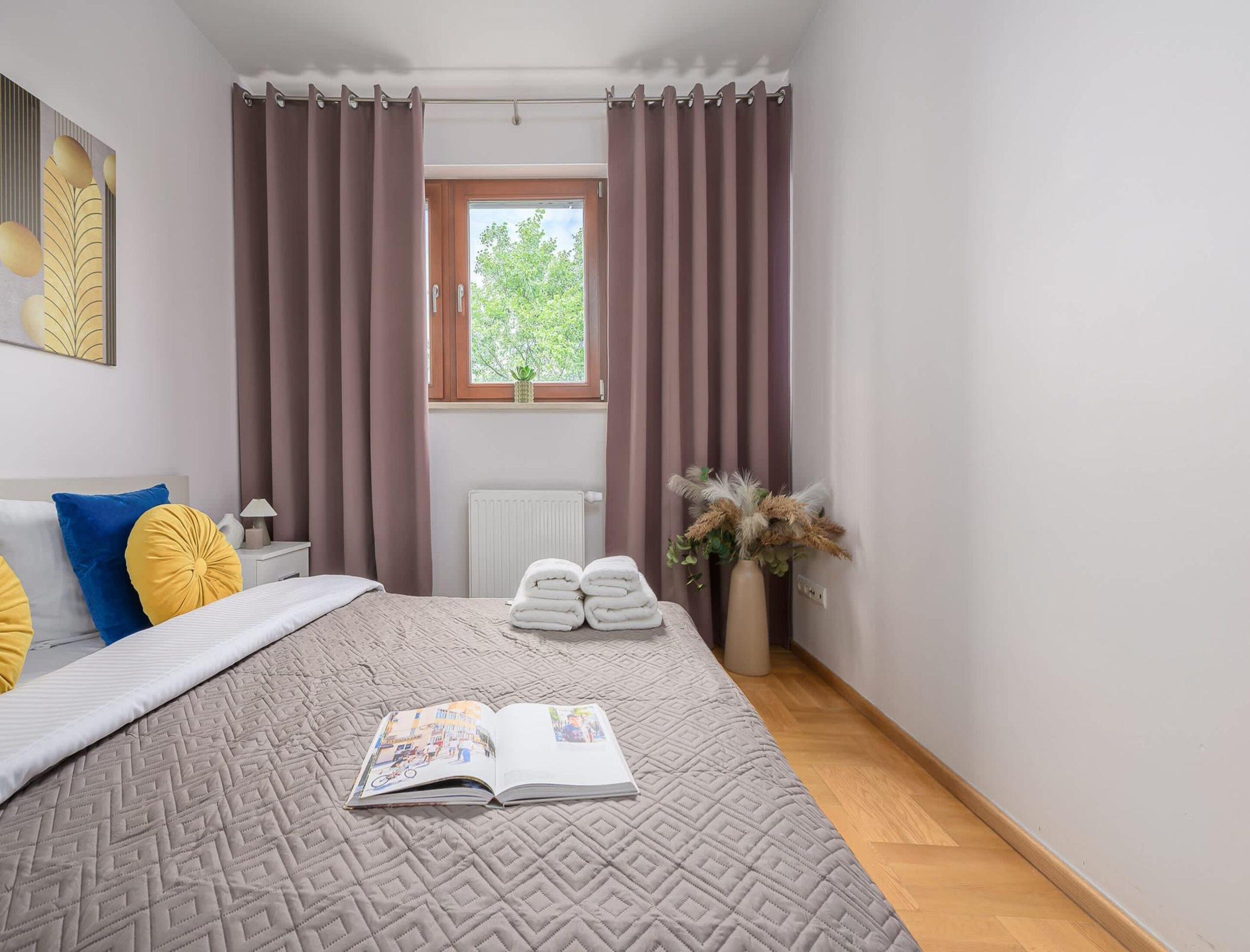 A bedroom with a bed, a window with curtains, a radiator, a bedside table with a lamp, and a book on the bed.