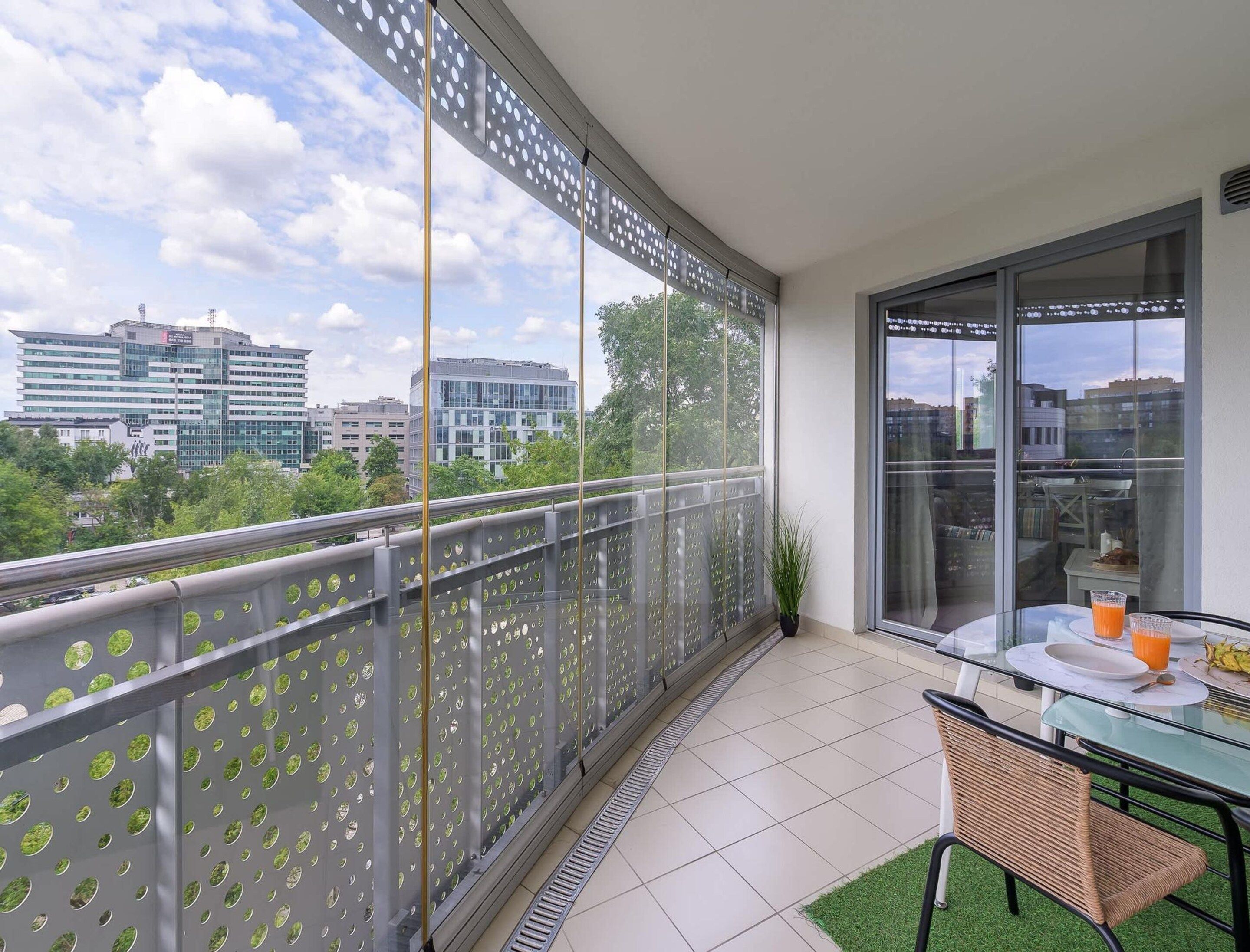 A balcony with a view of buildings, a glass table with a chair, and a potted plant.