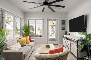 A modern living room with a sofa, coffee table, and TV. There are windows with blinds, a ceiling fan, and a decorative wall piece. - Bay Views & Walk to Little Italy - Prime Location (San Diego)