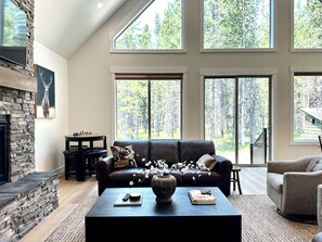 A spacious living room with a fireplace, a large window with a forest view, a leather sofa, and a coffee table with books and a vase. - Pleasant 3-bedroom cabin in Island Park for a relaxing getaway (Island Park)