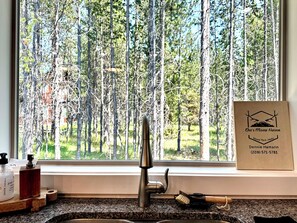 A kitchen with a window view of a forest, a stainless steel faucet, and a framed sign on the window sill. - Pleasant 3-bedroom cabin in Island Park for a relaxing getaway (Island Park)
