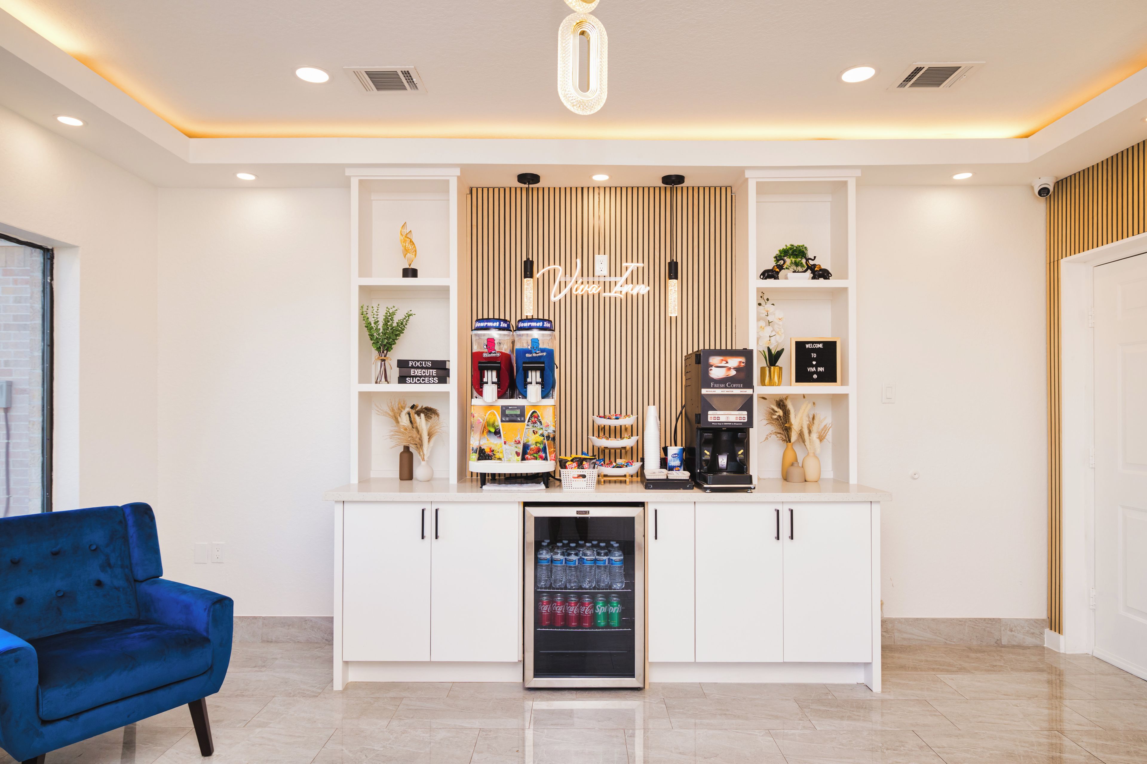 Una kitchenette moderna con una cafetera, un mini refrigerador lleno de bebidas y una planta decorativa.