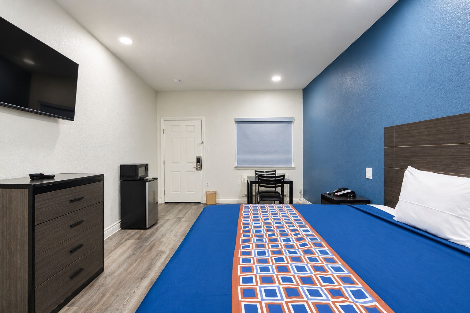 Habitación de hotel con cama, escritorio, silla, cómoda, televisor montado en la pared y una puerta.