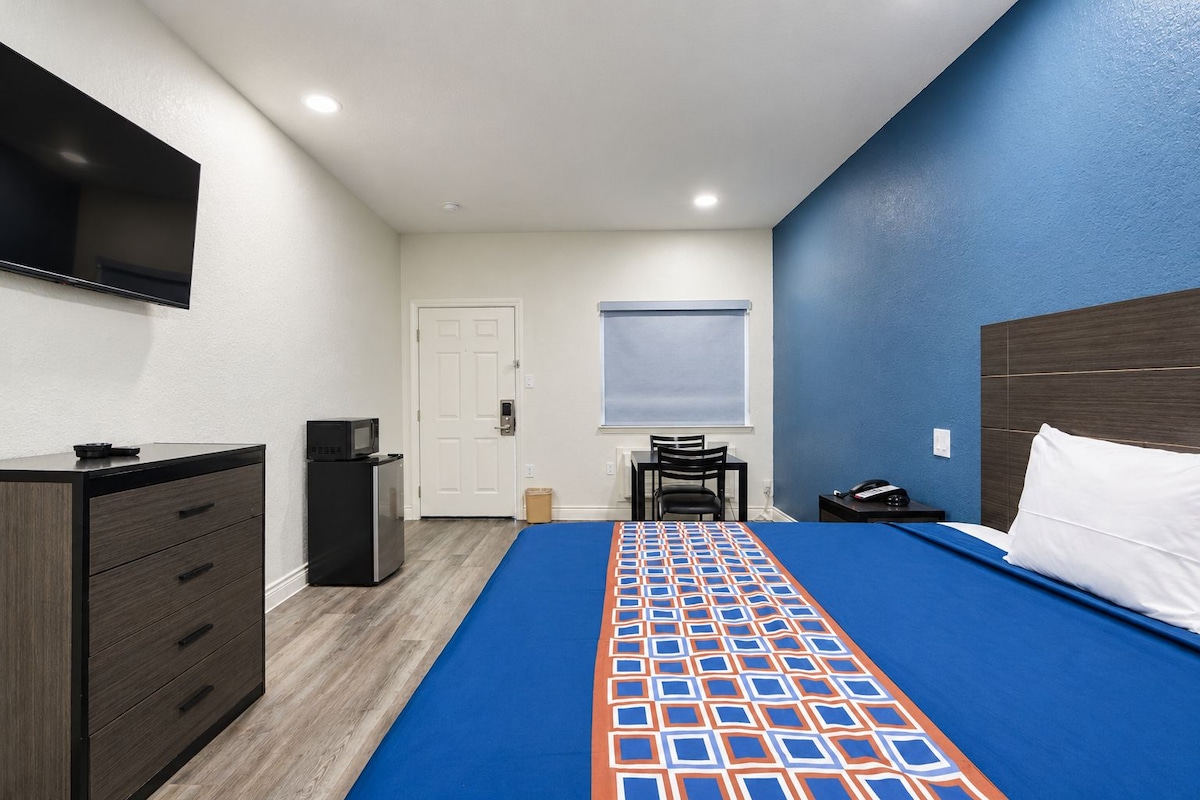 A hotel room with a bed, a desk, a chair, a dresser, a television mounted on the wall, and a door.