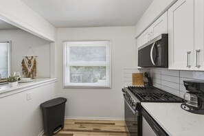 A compact kitchen with white cabinets, a stove, a microwave, and a window with blinds. - Cozy Stylish Apt in Omaha`s Medical District! (Omaha)