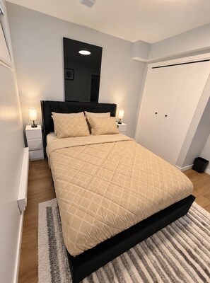 A bedroom with a bed, two bedside tables, a nightstand, a wall-mounted mirror, a ceiling light, and a door. - Cozy Private 1-Bedroom Suite |  Free Parking & Wi-Fi (Regina)