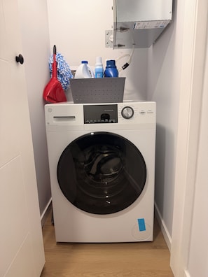 A front-loading washing machine with a control panel, a basket on top, and a mop and bucket to the left. - Cozy Private 1-Bedroom Suite |  Free Parking & Wi-Fi (Regina)