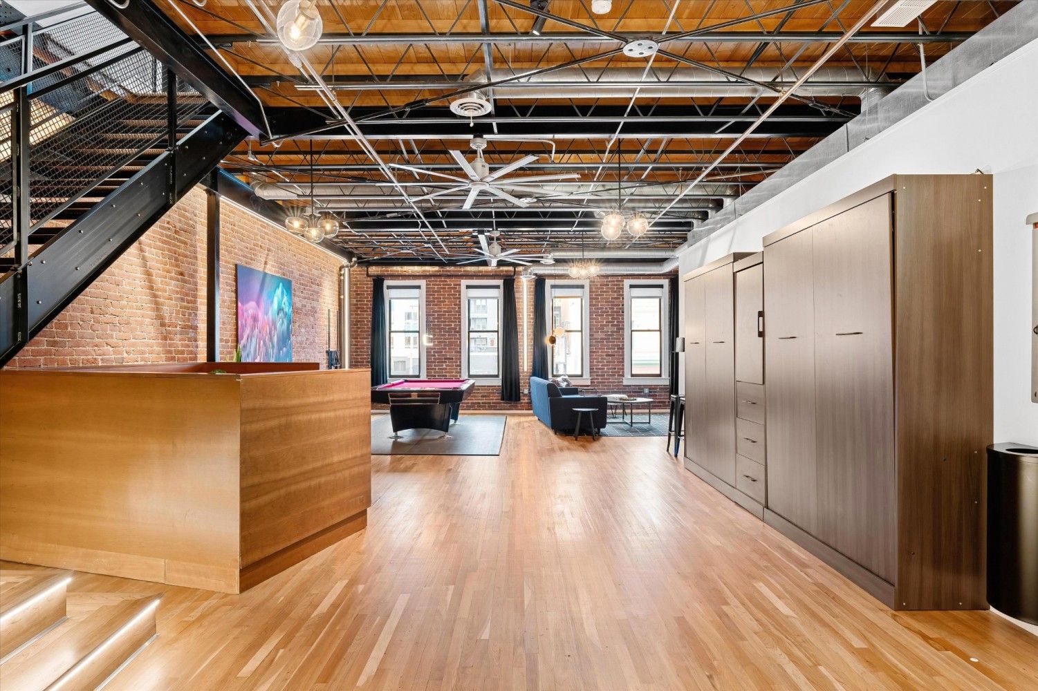 A spacious room with a wooden floor, a pool table, a sofa, and a staircase.