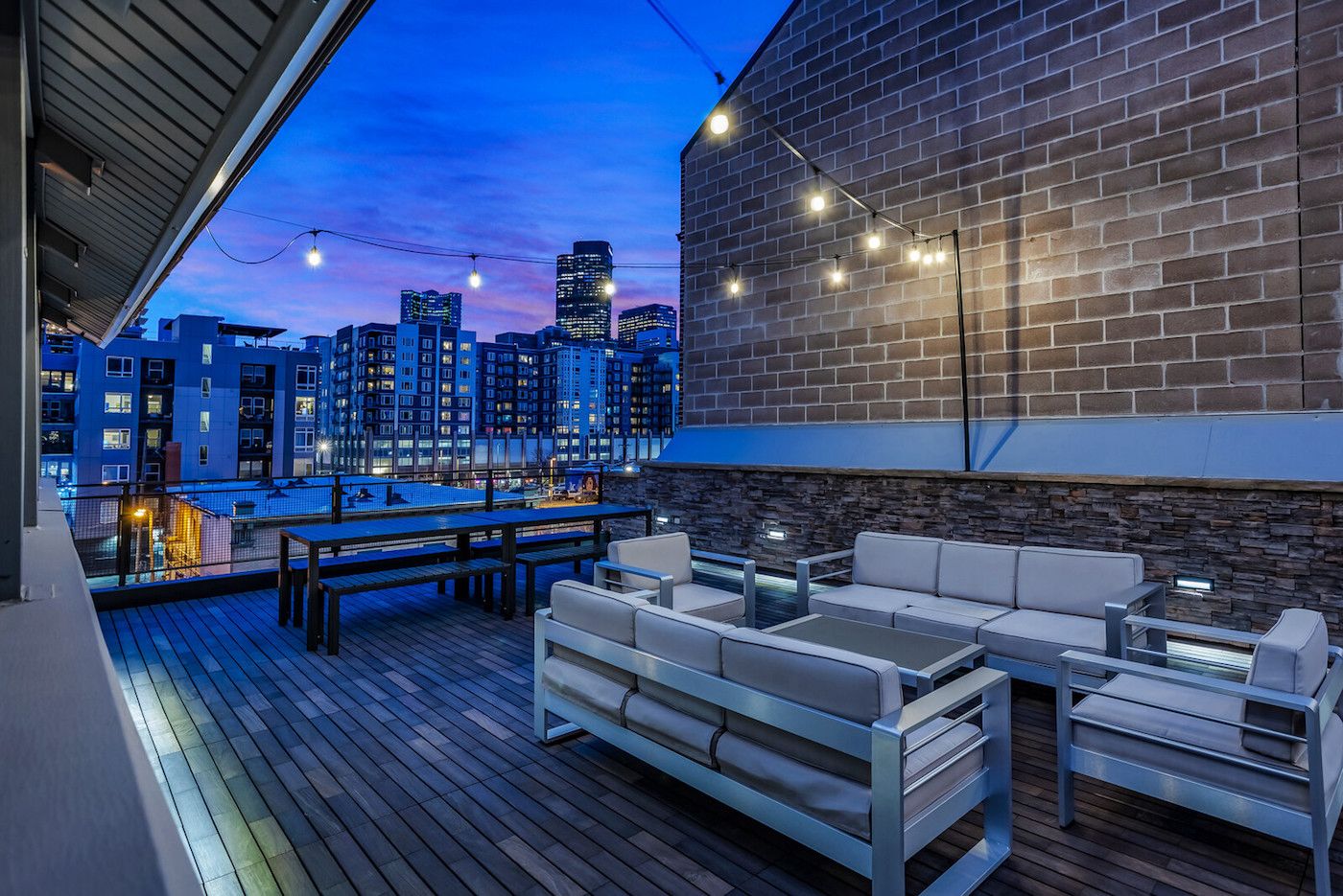An outdoor deck with a city view, featuring a brick wall, a seating area with cushions, and string lights.