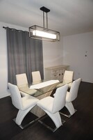 Comedor moderno con una mesa de vidrio y sillas blancas, suelo de madera oscura y un mueble claro al fondo.