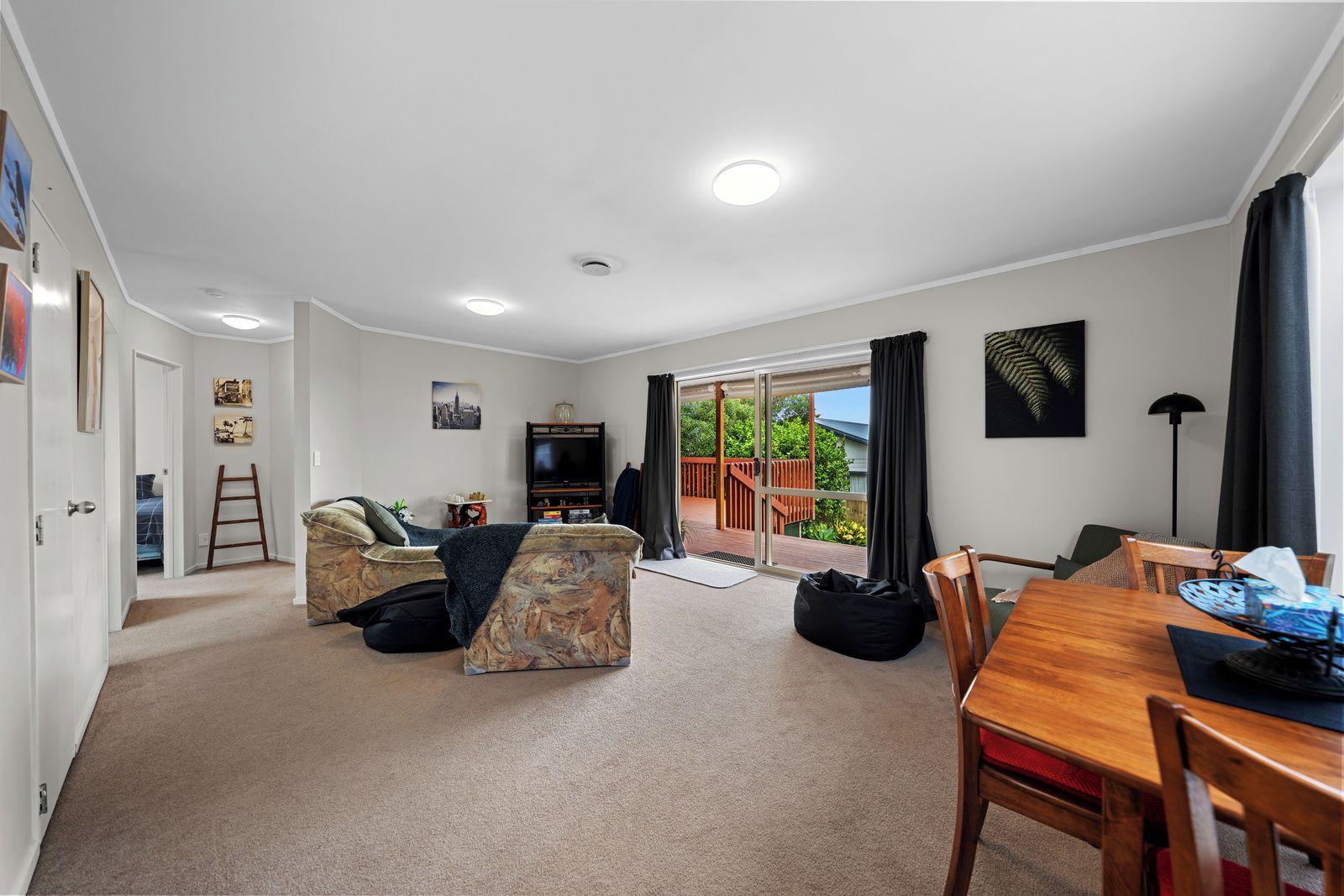 A living room with a sofa, a dining table, and a TV. There is a balcony with a view of greenery.