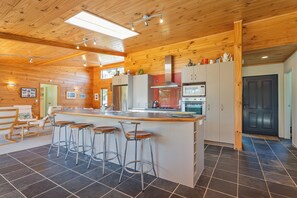 A kitchen with a central island, bar stools, stainless steel appliances, and wooden ceiling. - The Holiday Hideaway - Pauanui Holiday Home (Pauanui)