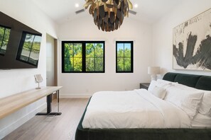 Ein modernes Schlafzimmer mit einem großen Bett, zwei Fenstern mit Blick auf Bäume, einem Schreibtisch mit Stuhl und einer außergewöhnlichen Kronleuchter.