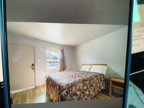 A bedroom with a bed, a window with curtains, a door, and a small wooden nightstand.