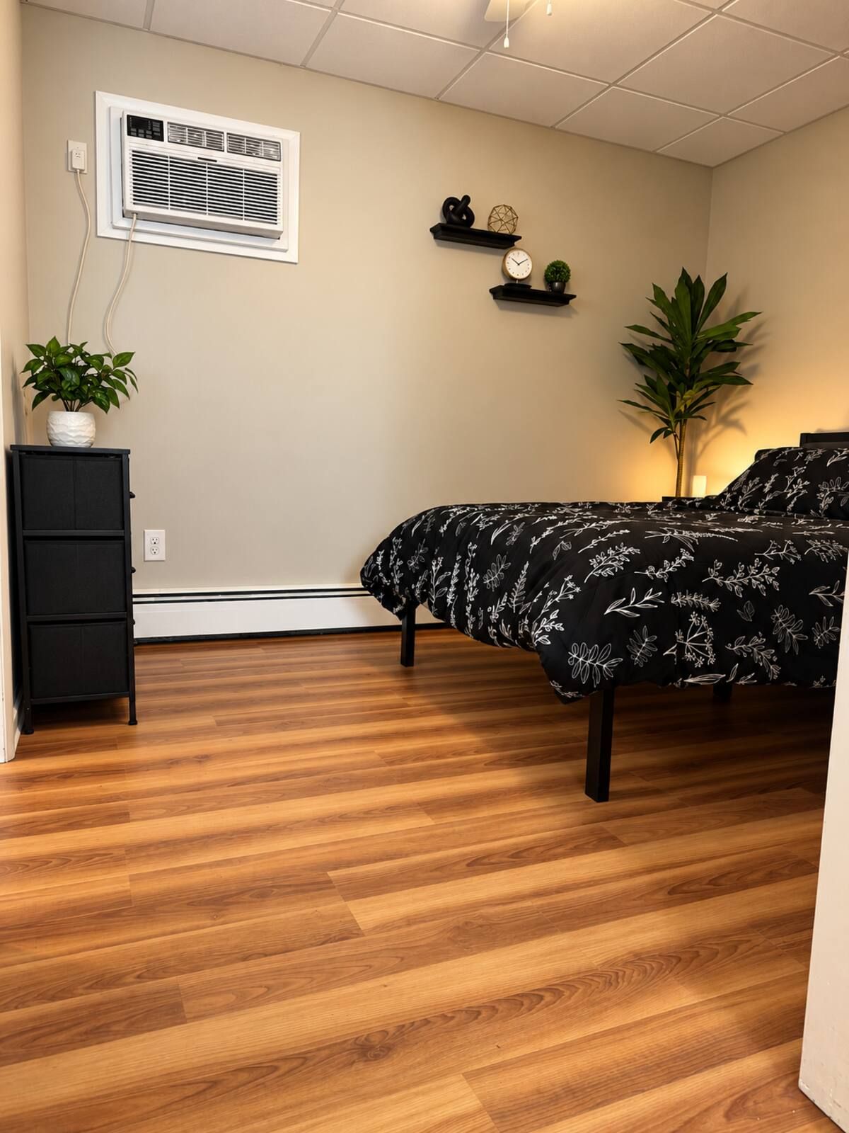 A bedroom with a bed, a dresser, a potted plant, a wall-mounted heater, and a shelf with decorative items.