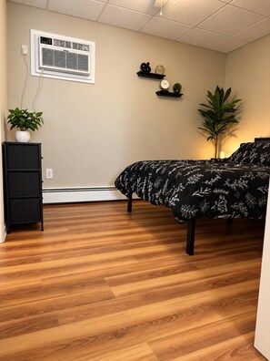 A bedroom with a bed, a dresser, a potted plant, a wall-mounted heater, and a shelf with decorative items. - Modern 1BR Cottage Retreat Near College (Swarthmore)