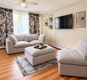A living room with a sectional sofa, an ottoman, a flat-screen TV, and decorative plants. - Modern 1BR Cottage Retreat Near College (Swarthmore)
