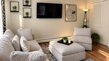 A living room with a sofa, armchair, ottoman, flat-screen TV, artwork, floor lamp, and a small table with candles.