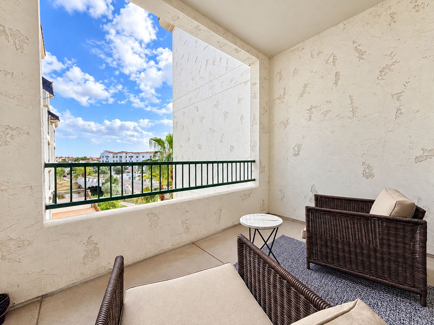 A balcony with a view of buildings and palm trees, furnished with wicker furniture and a small round table.