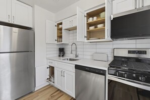 A modern kitchen with white cabinetry, a stainless steel refrigerator, a dishwasher, a coffee maker, a toaster, and a black stove. - Cozy Stylish Apt in Omaha`s Medical District! (Omaha)