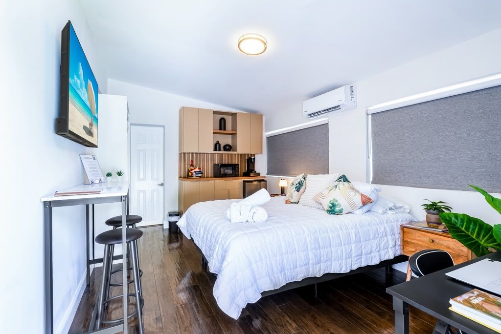 A compact hotel room with a bed, a desk, a chair, a small kitchenette, and a large window.