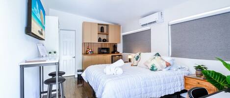 A compact hotel room with a bed, a desk, a chair, a small kitchenette, and a large window.