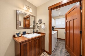 Une salle de bain avec un meuble-lavabo en bois, un grand miroir, un évier et une porte donnant sur une buanderie.