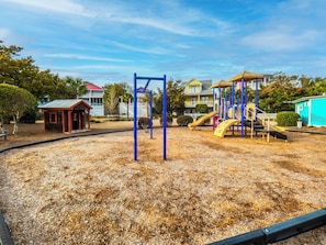 A playground with swings and slides, surrounded by a sandy area and residential buildings in the background. - Seaglass Grove (Kure Beach)