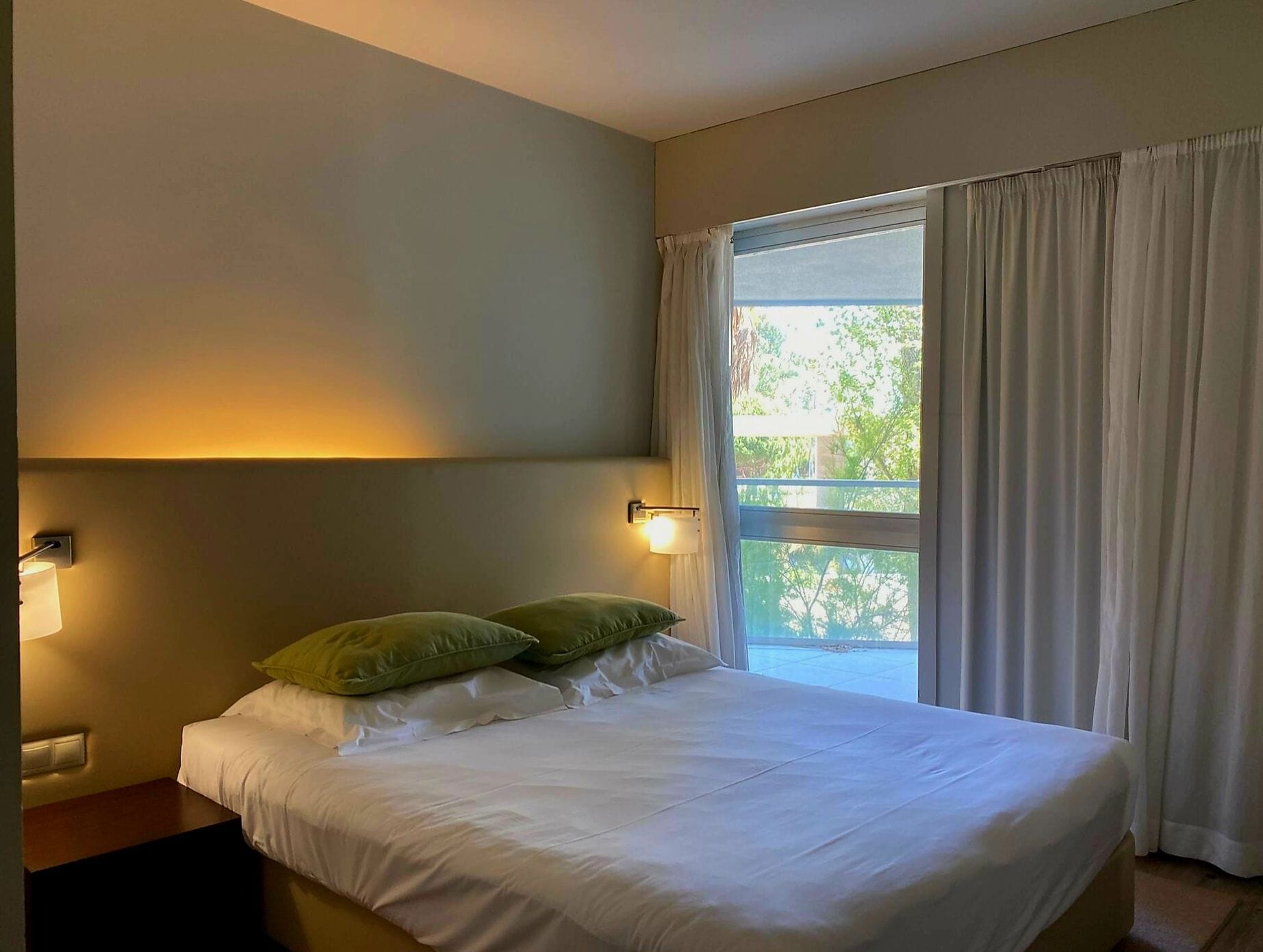 A hotel room with a bed, two pillows, a nightstand, a lamp, and a window with curtains.