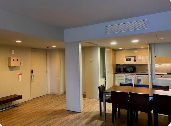 A compact kitchen with a microwave, toaster, and refrigerator. There is a dining table with chairs and a bench. The room has a wooden floor and white walls.