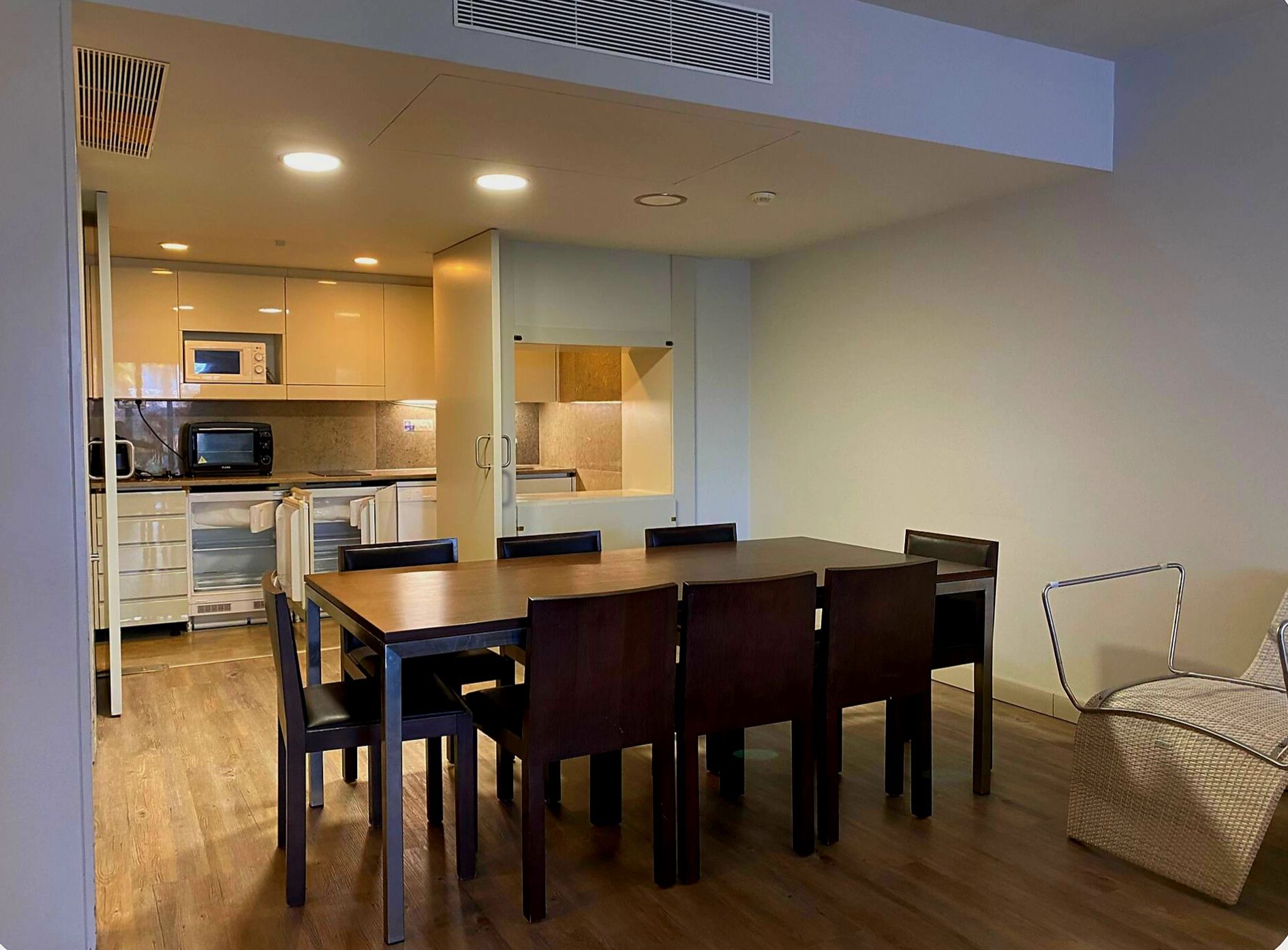 A compact kitchen with modern appliances, a dining area with a wooden table and chairs, and a small seating area with a woven chair.