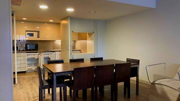 A compact kitchen with modern appliances, a dining area with a wooden table and chairs, and a small seating area with a woven chair.