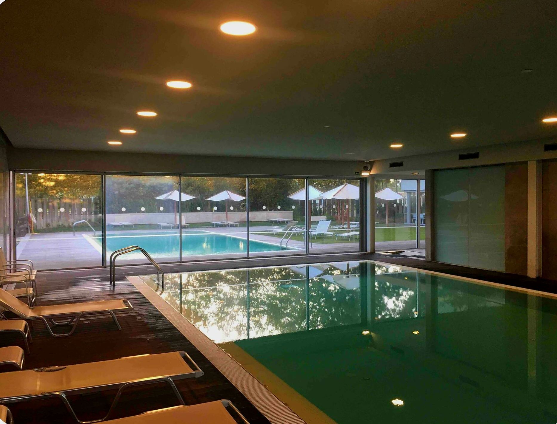 Une piscine intérieure avec des parois en verre transparent, des chaises longues et une vue sur l’extérieur.