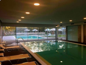 An indoor swimming pool with clear glass walls, lounge chairs, and a view of the outside.