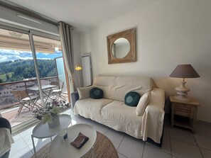 A living room with a sofa, a round table, a lamp, a mirror, and a view of a mountainous landscape through large windows. - Bel appartement avec terrasse et garage - vue pistes (Péone)