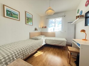 Une petite pièce bien éclairée, avec deux lits simples, un bureau, une chaise, une télévision et une fenêtre donnant sur le ciel.
