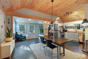 Eine moderne Küche mit Holzbalkendecke, ein Essbereich mit Tisch und Stühlen sowie ein Wohnzimmer mit Sofa und Pflanze.