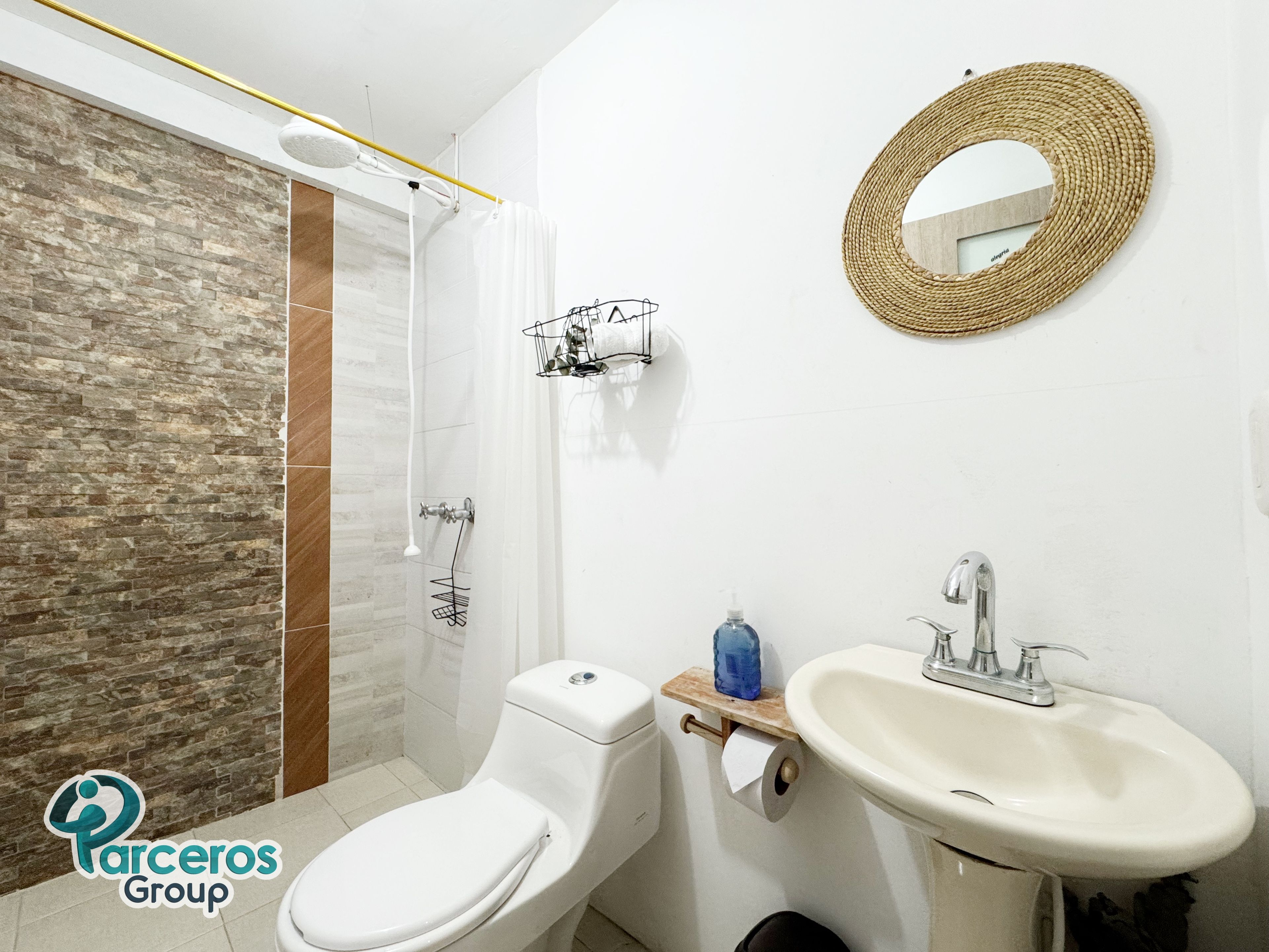 A bathroom with a shower, toilet, sink, and a woven mirror.