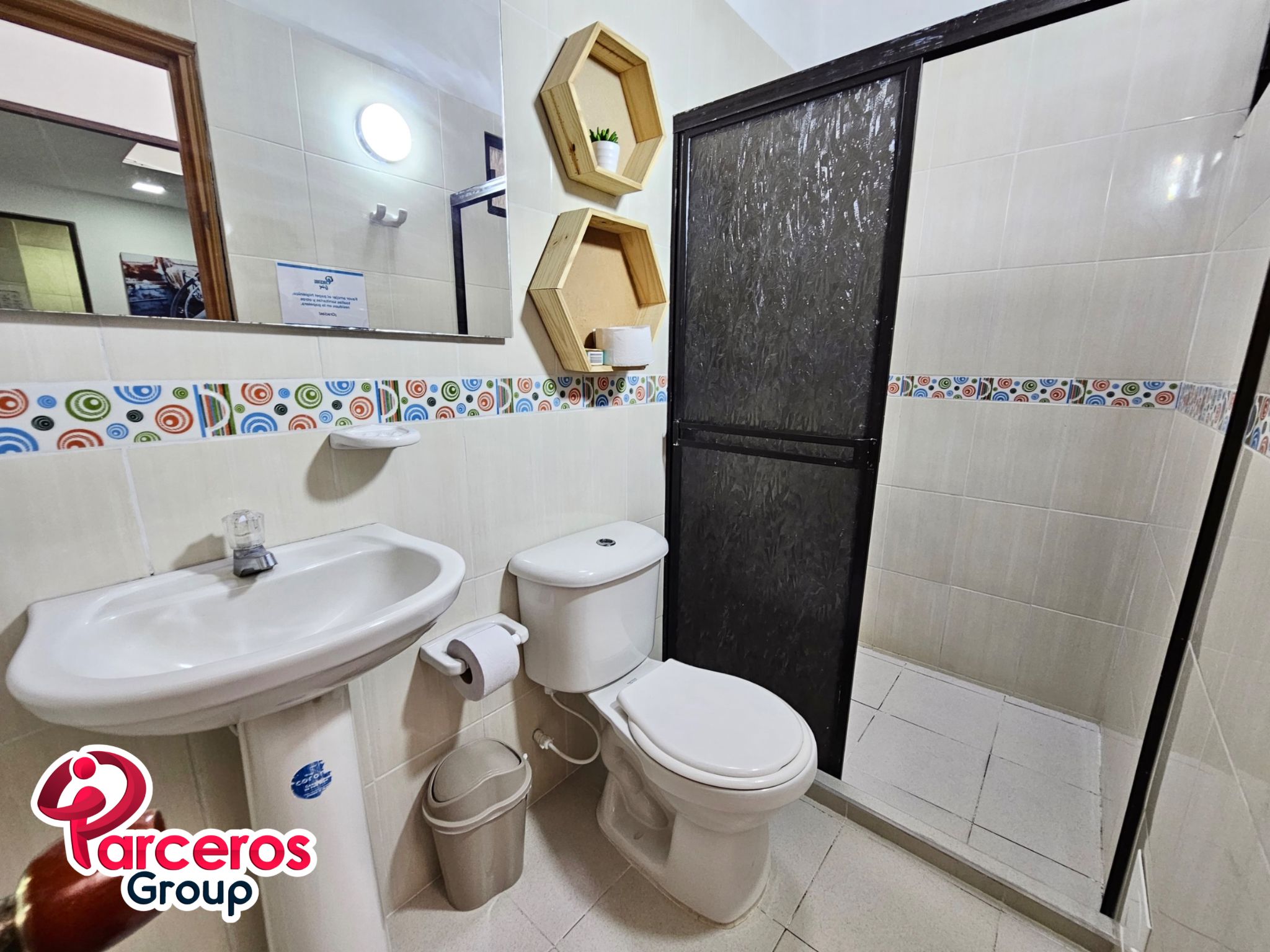 Une salle de bain équipée d’un lavabo, de toilettes et d’un espace douche. On y trouve une poubelle et un miroir au-dessus du lavabo.
