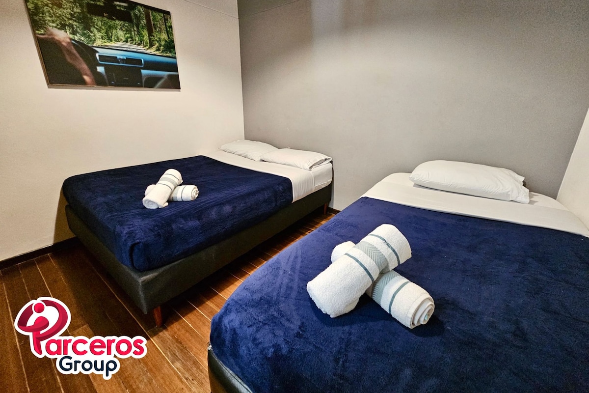 Habitación de hotel con dos camas, cada una con cubrecama azul y almohadas blancas. Encima de las camas hay un cuadro enmarcado en la pared.