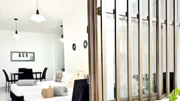 A modern hotel room with a bed, a desk, and a chair. There is a large window with vertical blinds, and a potted plant on the windowsill.