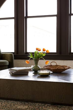 Ein Zimmer mit großem Fenster, einem Holztisch mit Blumenvase, einer Kerze und einem gewebten Korb.