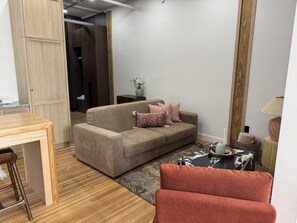 Ein eingerichtetes Wohnzimmer mit einer Couch, einem Couchtisch und einem Sessel. Im Vordergrund steht ein Holztisch und ein Barhocker.