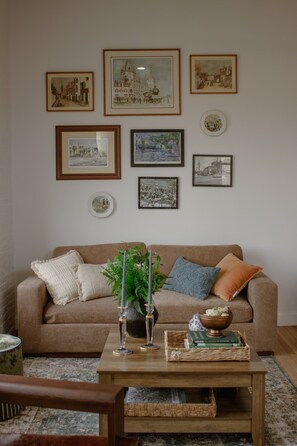 A living room with a brown sofa, framed artwork on the wall, a coffee table with decorative items, and a patterned rug. - Room 307, Ilium Luxury 2 Bd. (Troy)